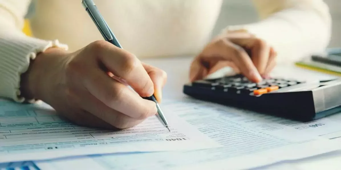 Manos escribiendo con lápiz sobre un formulario impreso mientras la otra mano marca teclas de una calculadora de escritorio.