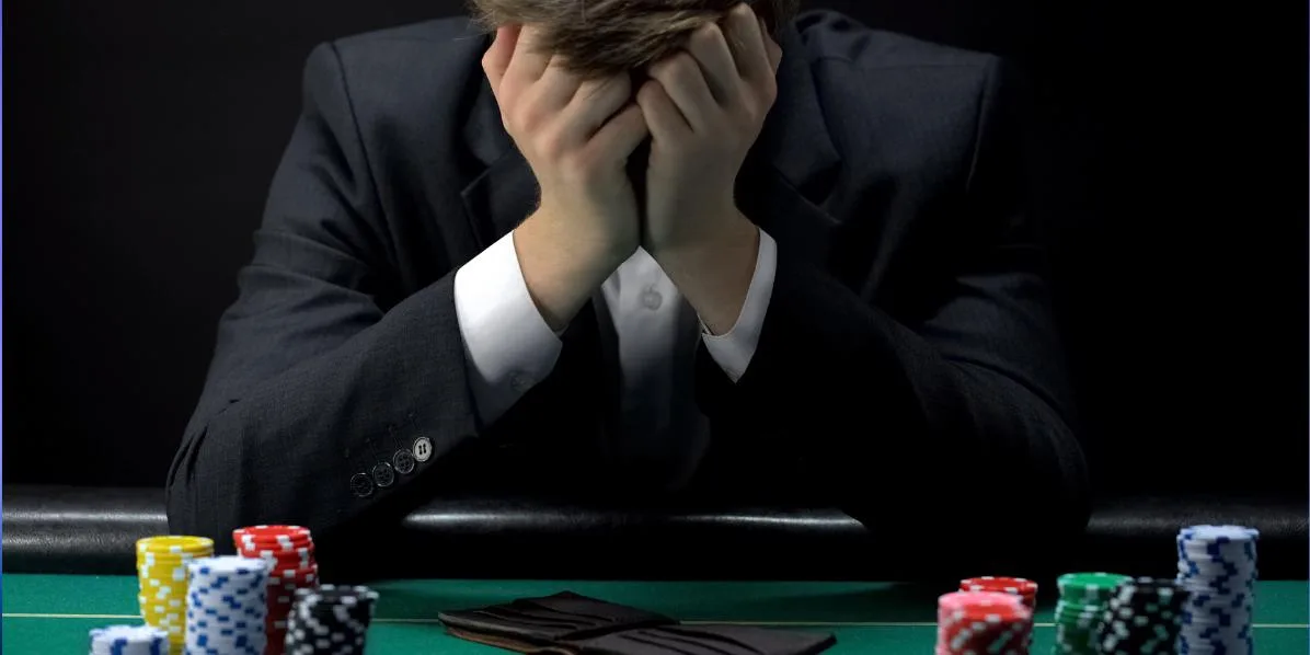 Hombre de traje sentado ante una mesa de paño verde con las manos cubriéndose la cara y fichas apiladas delante.